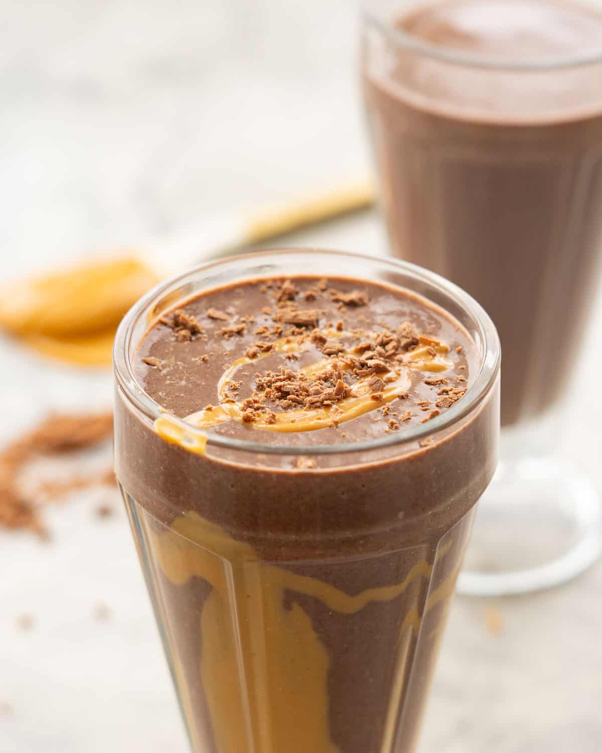 Chocolate peanut butter smoothie in tall smoothie glass poured to the rim, drizzled peanut butter on the inside of class and crumbled chocolate and peanut butter drizzled on top.