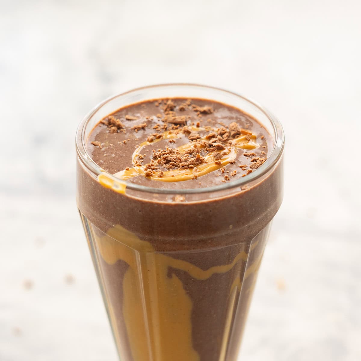 Chocolate peanut butter smoothie in tall smoothie glass poured to the rim, drizzled peanut butter on the inside of class and crumbled chocolate and peanut butter drizzled on top.