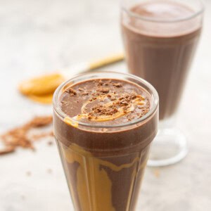 Chocolate peanut butter smoothie in tall smoothie glass poured to the rim, drizzled peanut butter on the inside of class and crumbled chocolate and peanut butter drizzled on top.