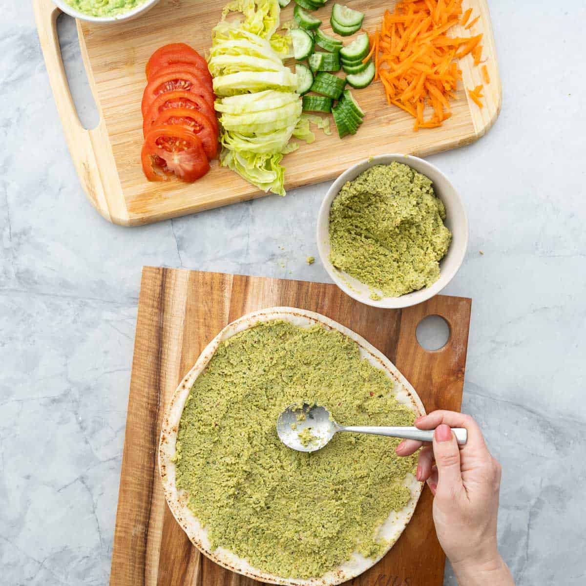 Falafel mix being spread evenly over pita bread with the back of a spoon. Fresh ingredients on wooden chopping board.