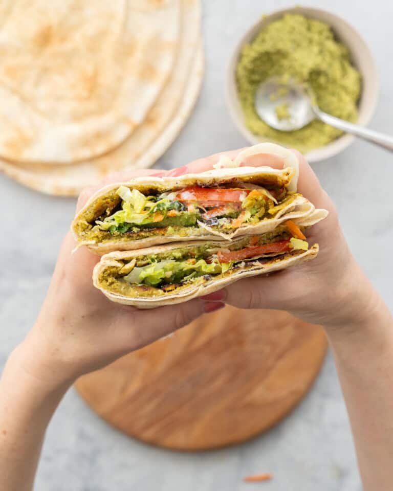 Falafel pita cut in half being held by a persons hands showing the layers of ingredients inside.