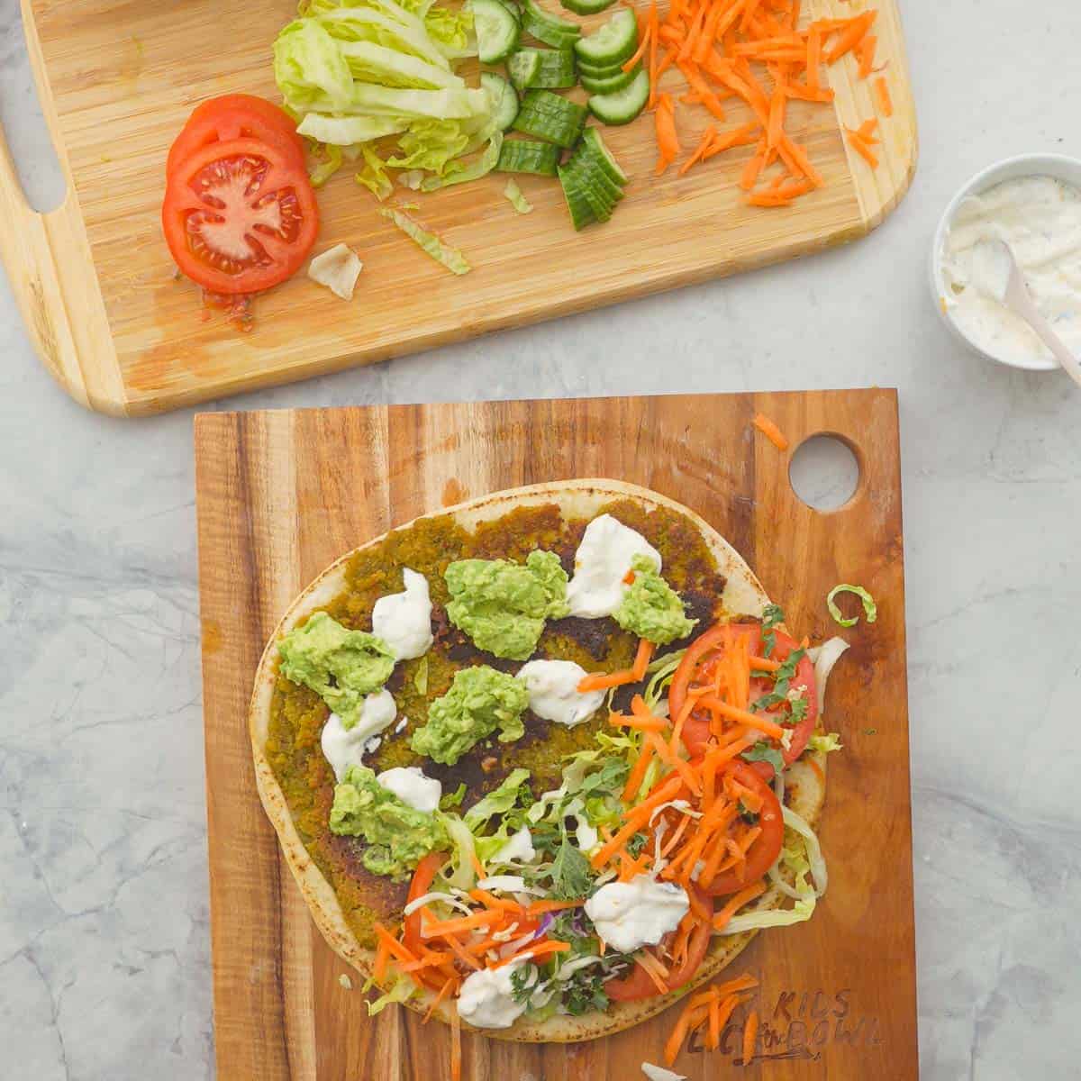 Pita Bread laid on wooden chopping board with Falafel mix spread on the bottom, avocado and feta dip spooned on top and fresh ingredients placed on one half.