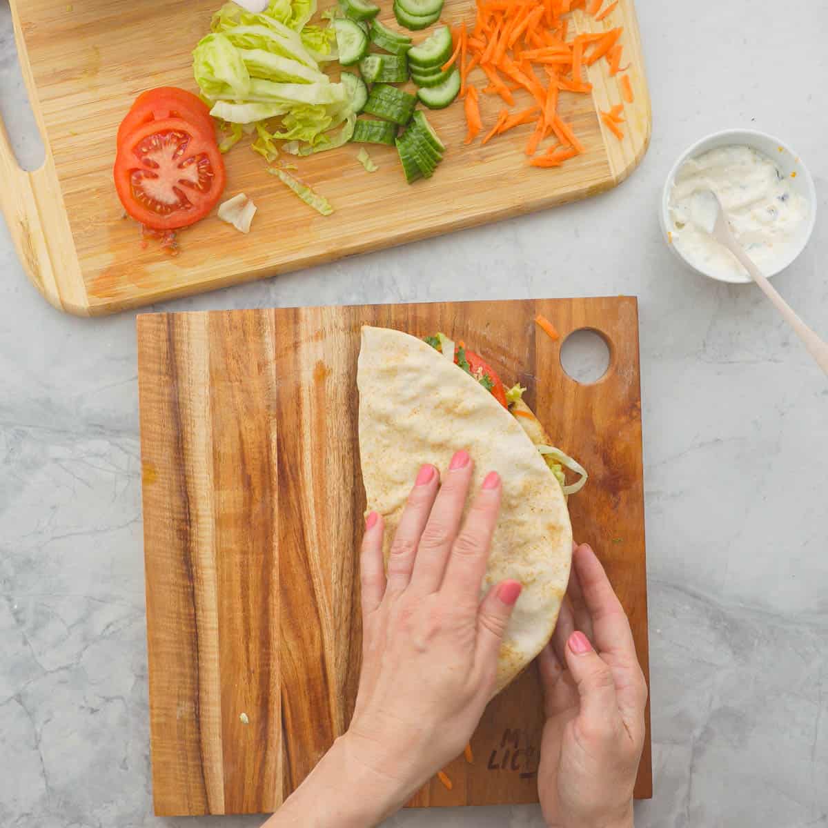 Falafel Pita bread being folded in half with fillings all inside. Wooden chopping board with left over fresh ingredients nearby.