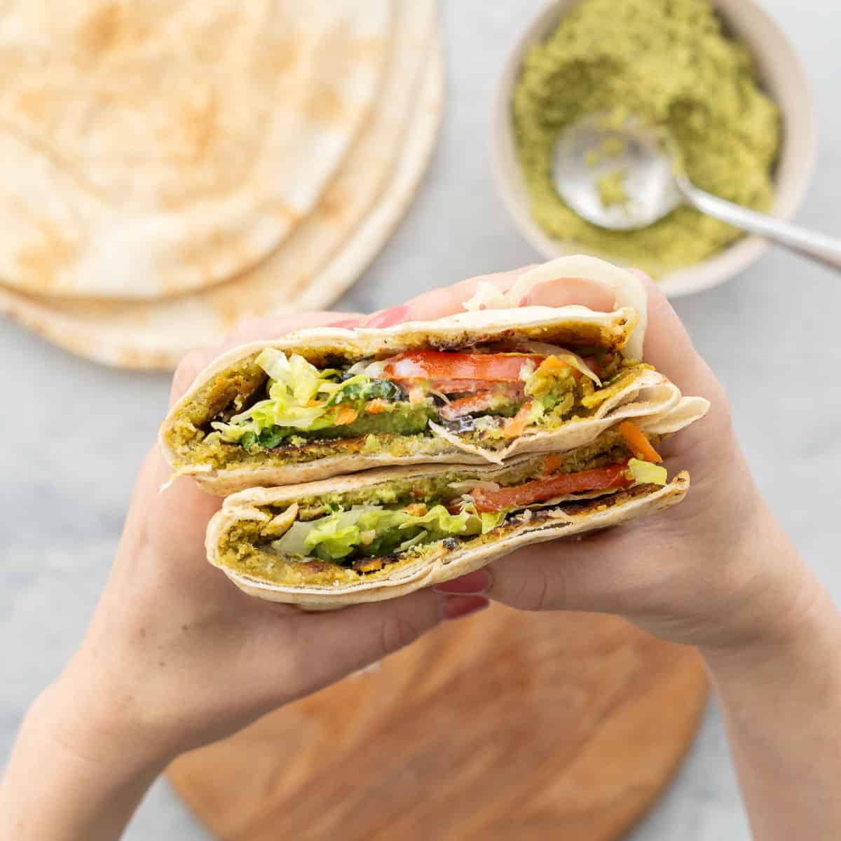 Falafel pita cut in half being held by a persons hands showing the layers of ingredients inside.