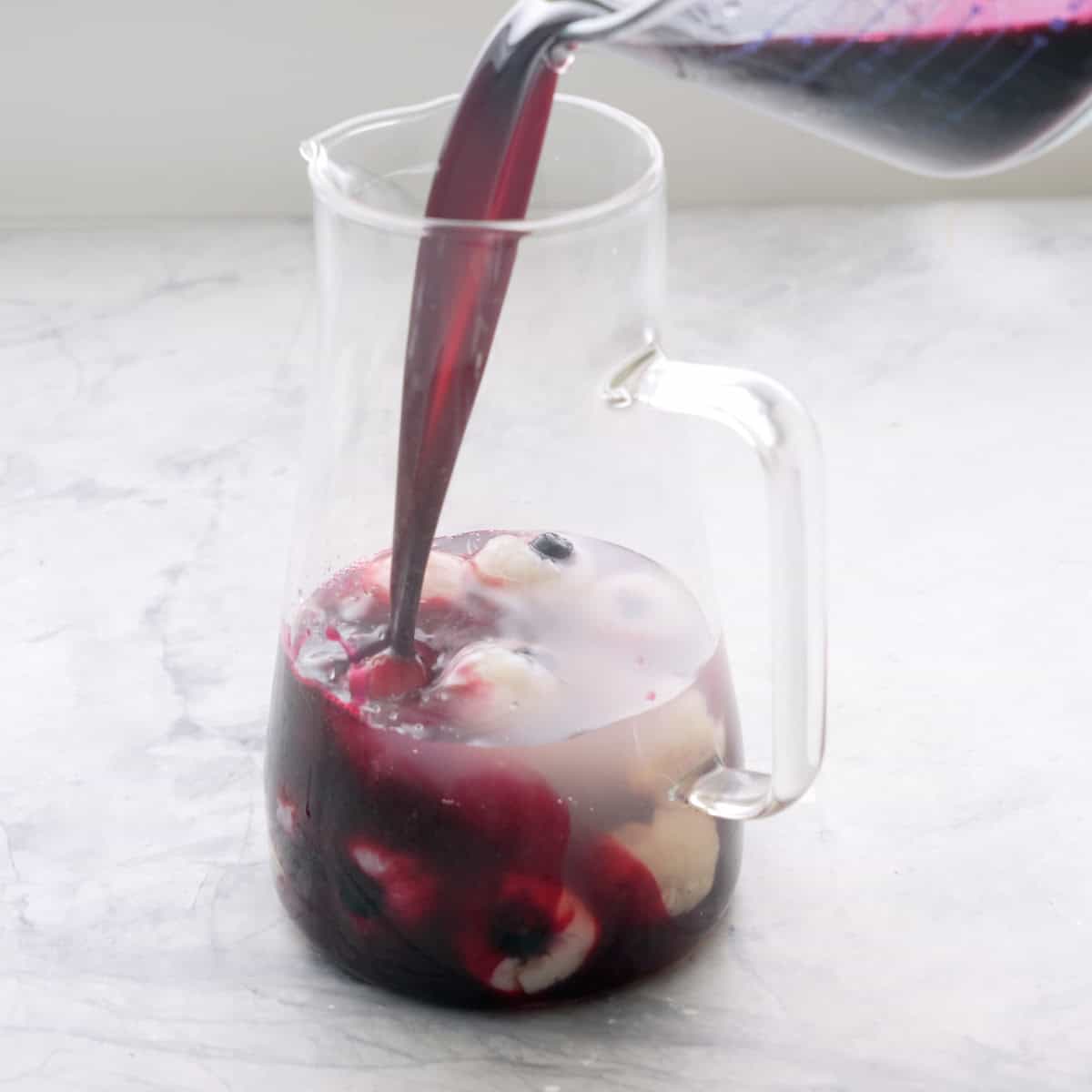 Punch being poured into glass jug ontop of lychee and blueberry eyeballs.