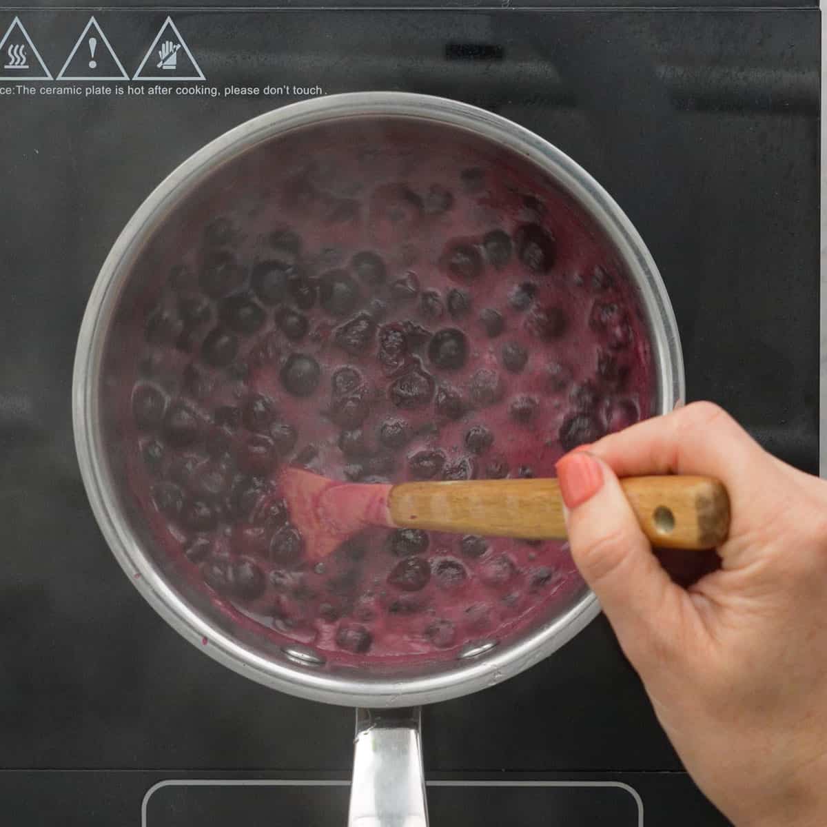 Frozen blueberries, honey and water in saucepan being stirred with spatula.