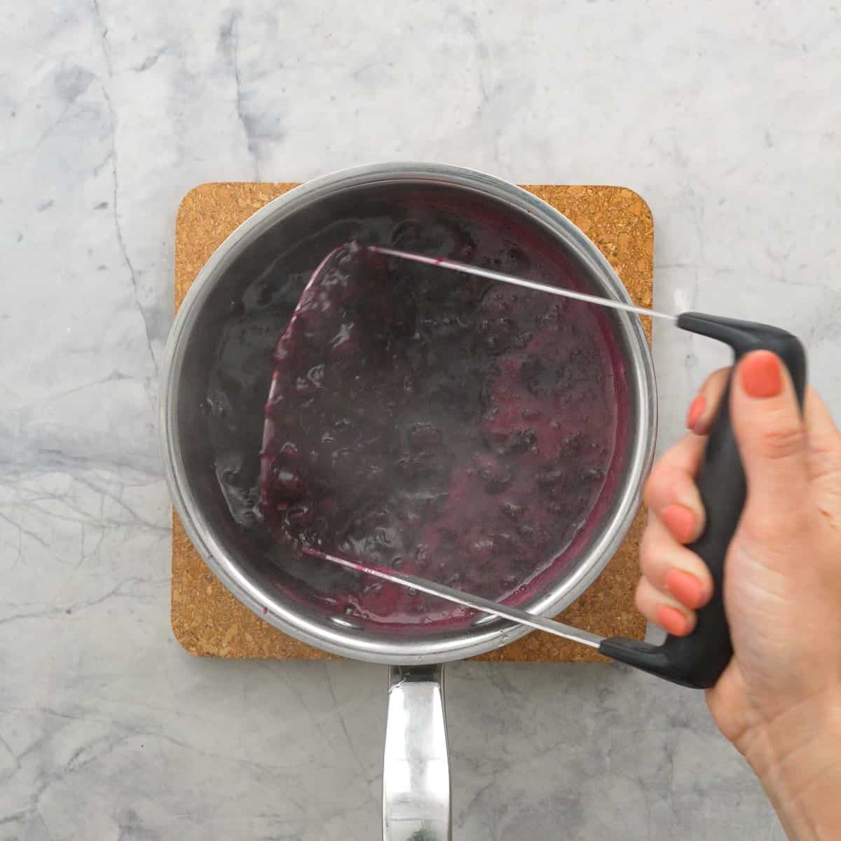 Hand holding a masher inside the pot of melted Blueberry mixture.