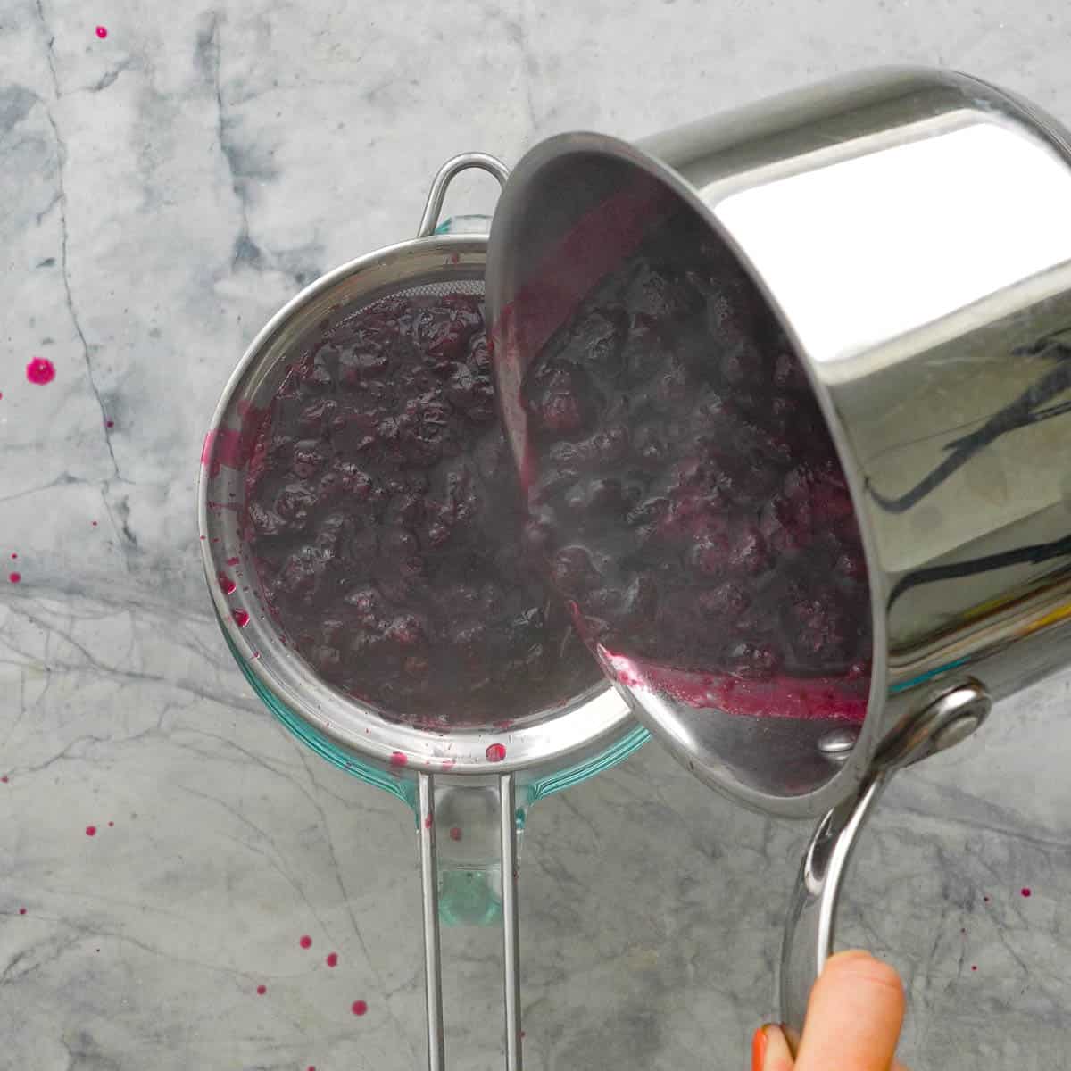 Pot pouring out mashed blueberry mixture into a sieve with bowl underneath catching the juice.