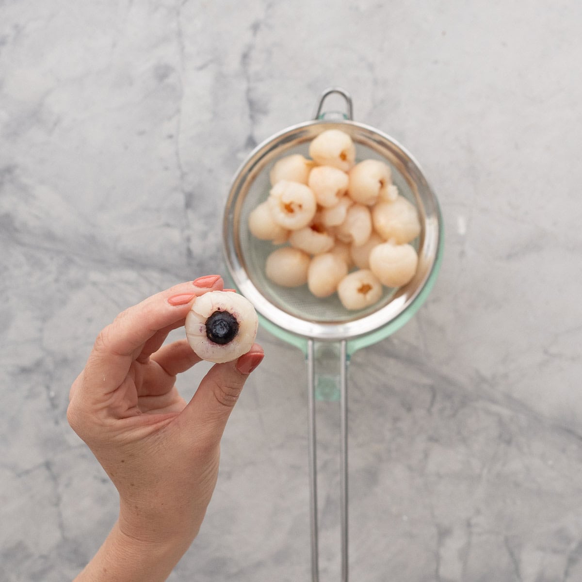 Lychees being strained on benchtop with hand holding a lychee with blueberry stuffed inside.