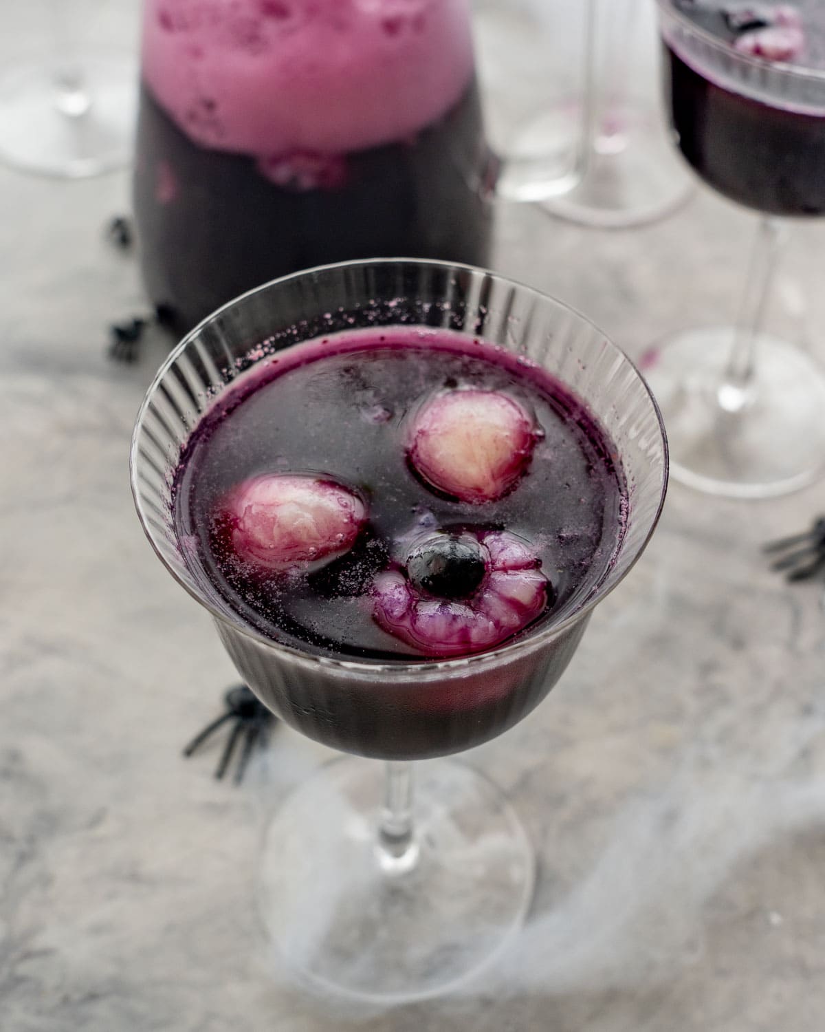 Halloween punch in tall glass, lychee and blueberry looking eyeballs floating on the top of the punch. Fake spiders and cobweb on benchtop.