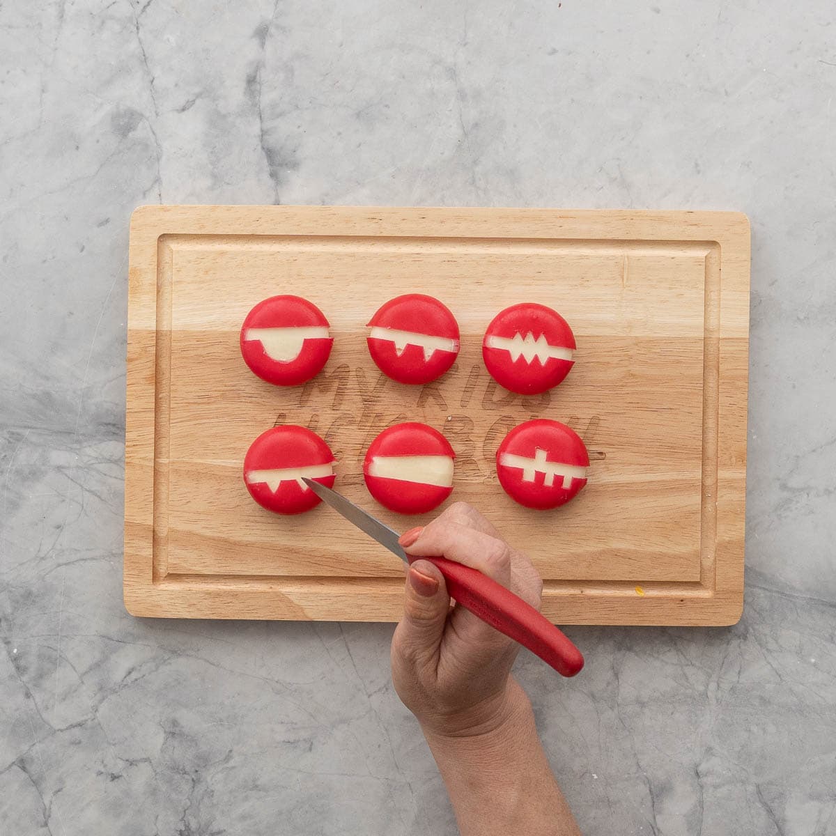 Six babybel cheese on wooden chopping board with the middle wax strip removed, hand using a small pointed sharp knife to create some fang shapes.