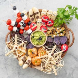 Halloween snack board on benchtop with many different halloween themed food. Cheese Monster, Mummy Cookies, Ants on a log, Spiderweb Pretzels, Fruit skewers, Banana Ghosts, Mandarin pumpkins, Guacamole and purple chips.