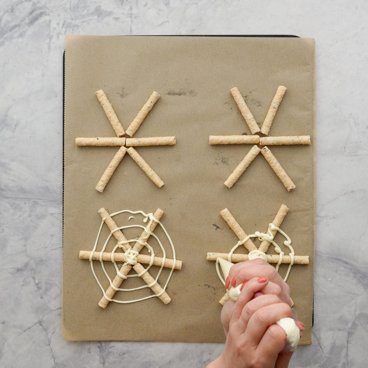 Pretzel sticks placed into 6 pronged star shape, hand holding a piping bag with melted white chocolate inside, drizzling in a circle on top of pretzels to create spiderwebs.