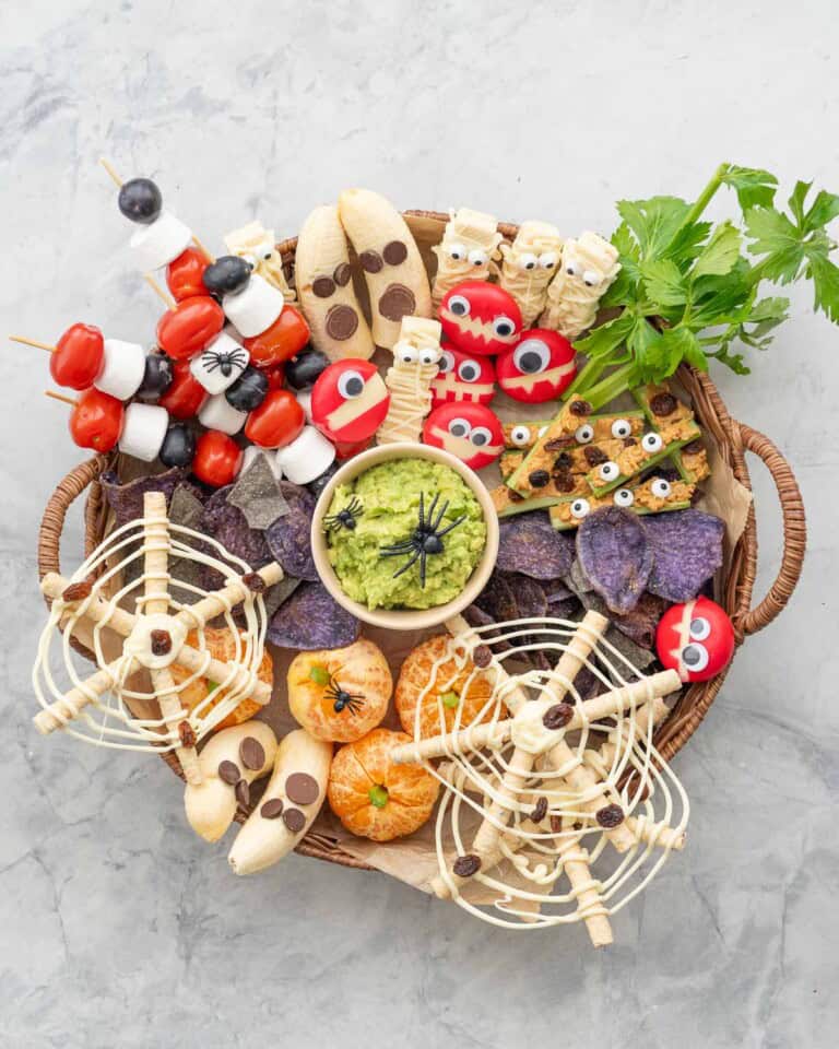 Halloween snack board on benchtop with many different halloween themed food. Cheese Monster, Mummy Cookies, Ants on a log, Spiderweb Pretzels, Fruit skewers, Banana Ghosts, Mandarin pumpkins, Guacamole and purple chips.