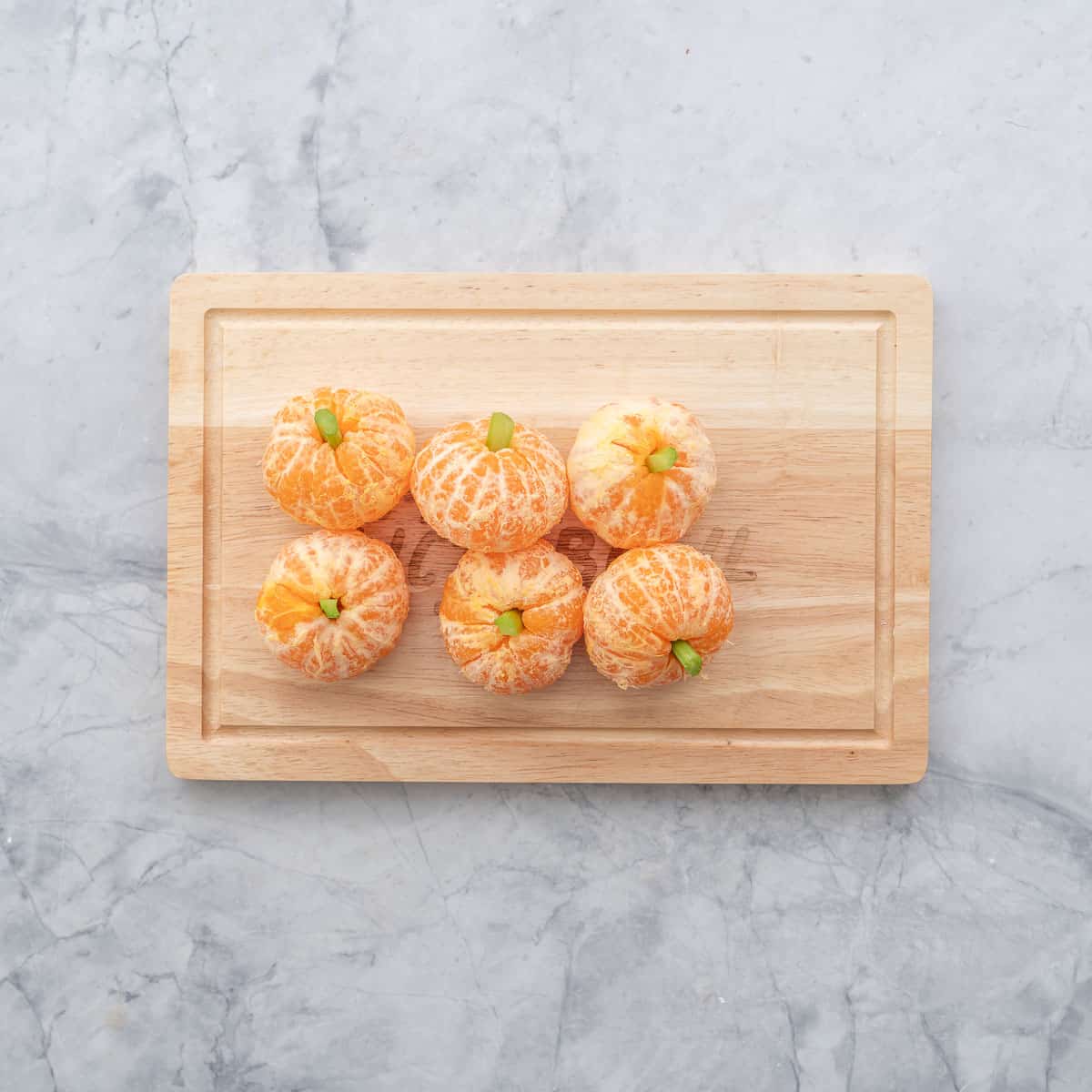 Six peeled mandarins on a chopping board with celery sticks as stalks so they look like pumpkins. 