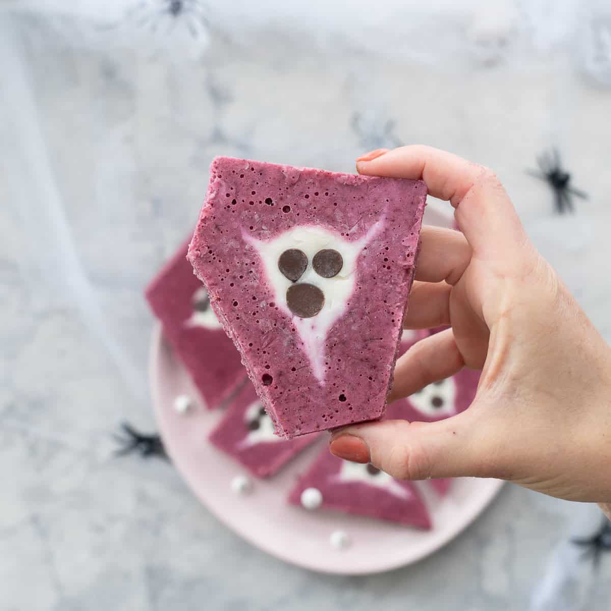 Halloween yogurt bark piece held up by persons hand, plate with the rest of yogurt bark blurred in background.