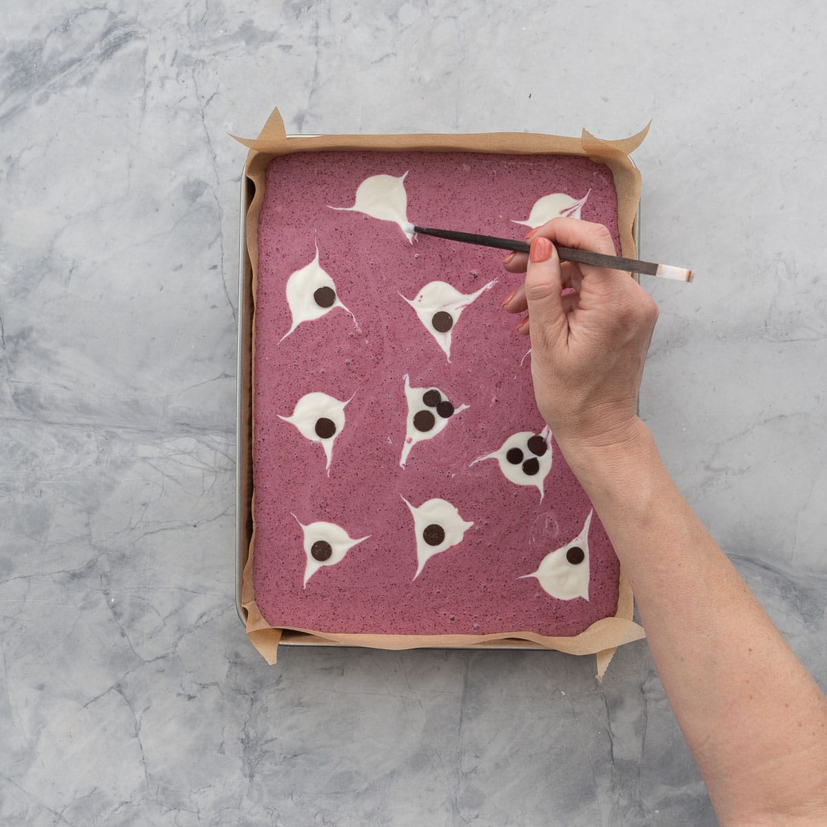 Berry yogurt mixture in lined baking tray with blobs of plain yogurt scooped on top, hand holding a chopstick swirling the yogurt to create arms and a tail of a ghost. Chocolate chips placed on as ghosts eyes and mouth.