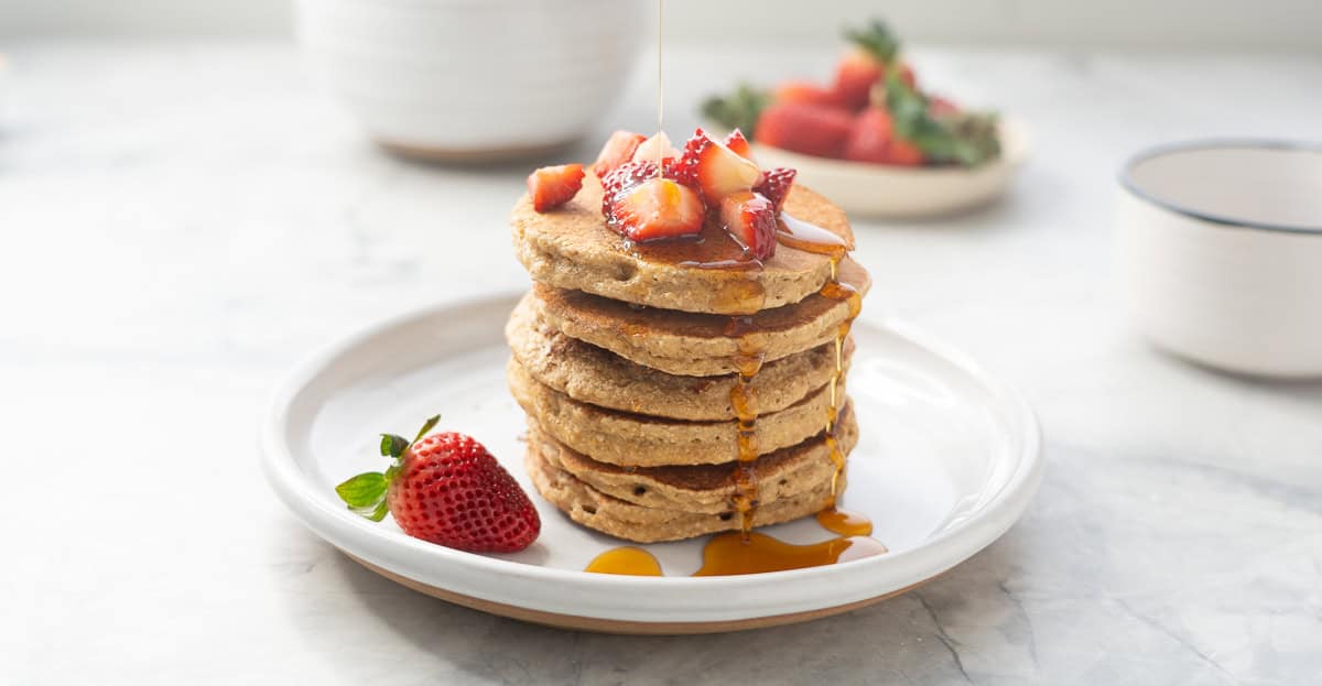 A stack of five oat flour pancakes with cut up strawberries on top and maple syrup drizzled over the top.