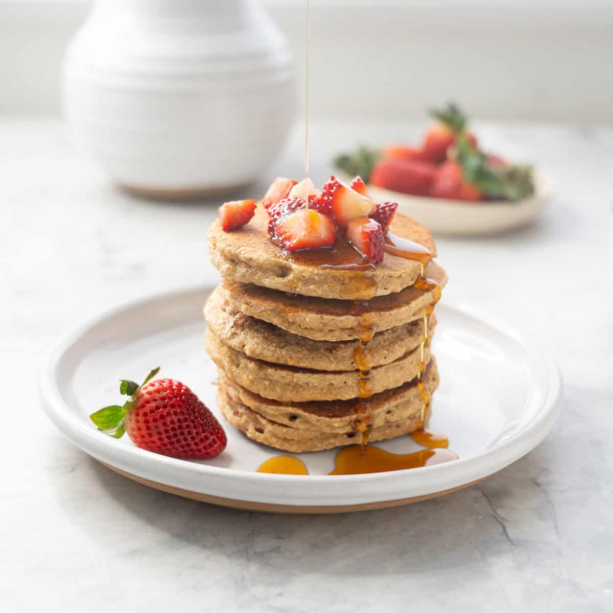 A stack of five oat flour pancakes with cut up strawberries on top and maple syrup drizzled over the top.