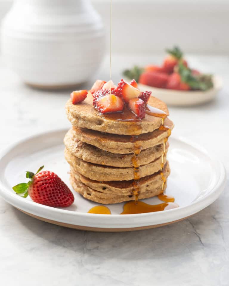 A stack of five oat flour pancakes with cut up strawberries on top and maple syrup drizzled over the top.