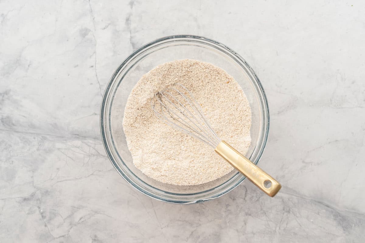 Dry ingredients for oat flour pancakes in glass mixing bowl on bench top with whisk inside.