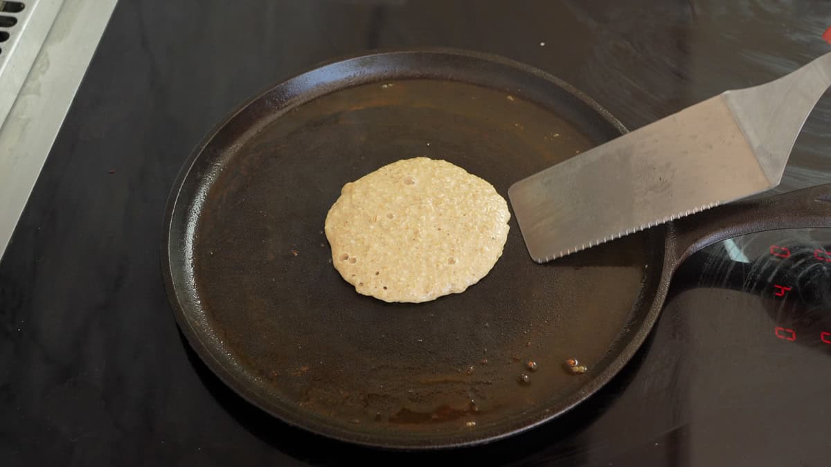 Oat flour pancake on a flat pan with bubbles forming on the top and a metal spatula about to flip it.