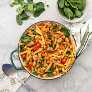 Sausage pasta in cast iron pan placed on benchtop, fresh spinach in a bowl and leaves scattered on the table.