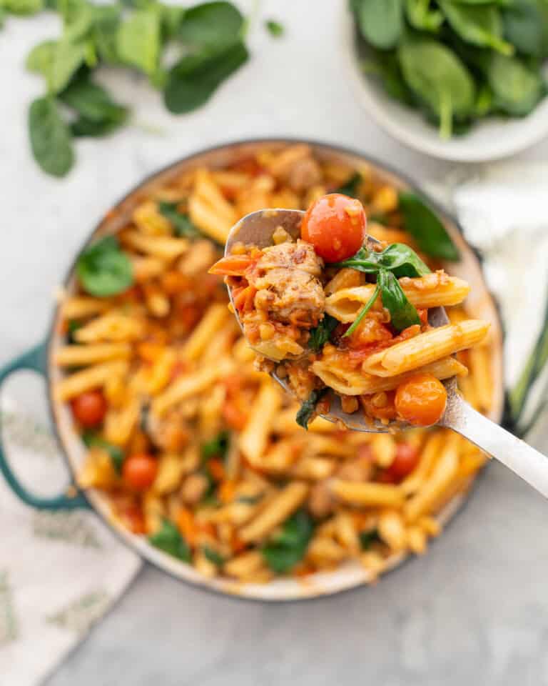 A big spoon holding up a scoop of sausage pasta with cast iron pan blurred in background with remainder of pasta.