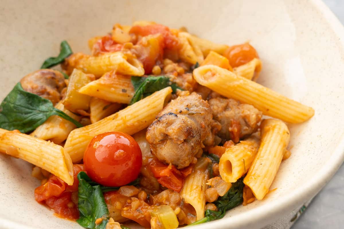 A bowl of Sausage pasta up close, cooked penne pasta, cherry tomatoes spinach, lentils and sausage.