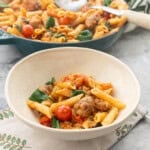 A bowl of served up sausage pasta with remainder of pasta in cast iron pan blurred in the background.