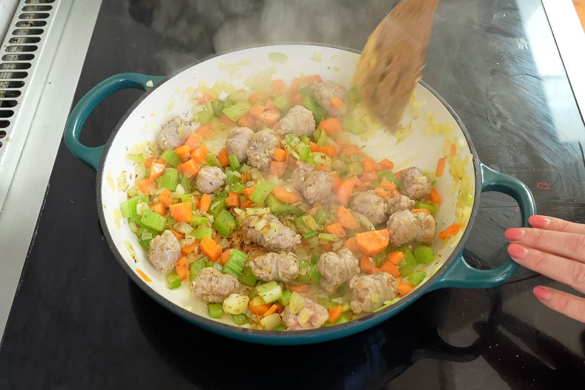 Celery, Carrot, onion and sausage being mixed with wooden spoon in cast iron pan on stove top.