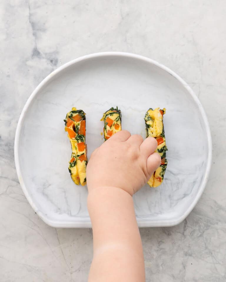 Three spinach omelette fingers on a child's silicone plate with a toddlers hand taking one of the fingers.