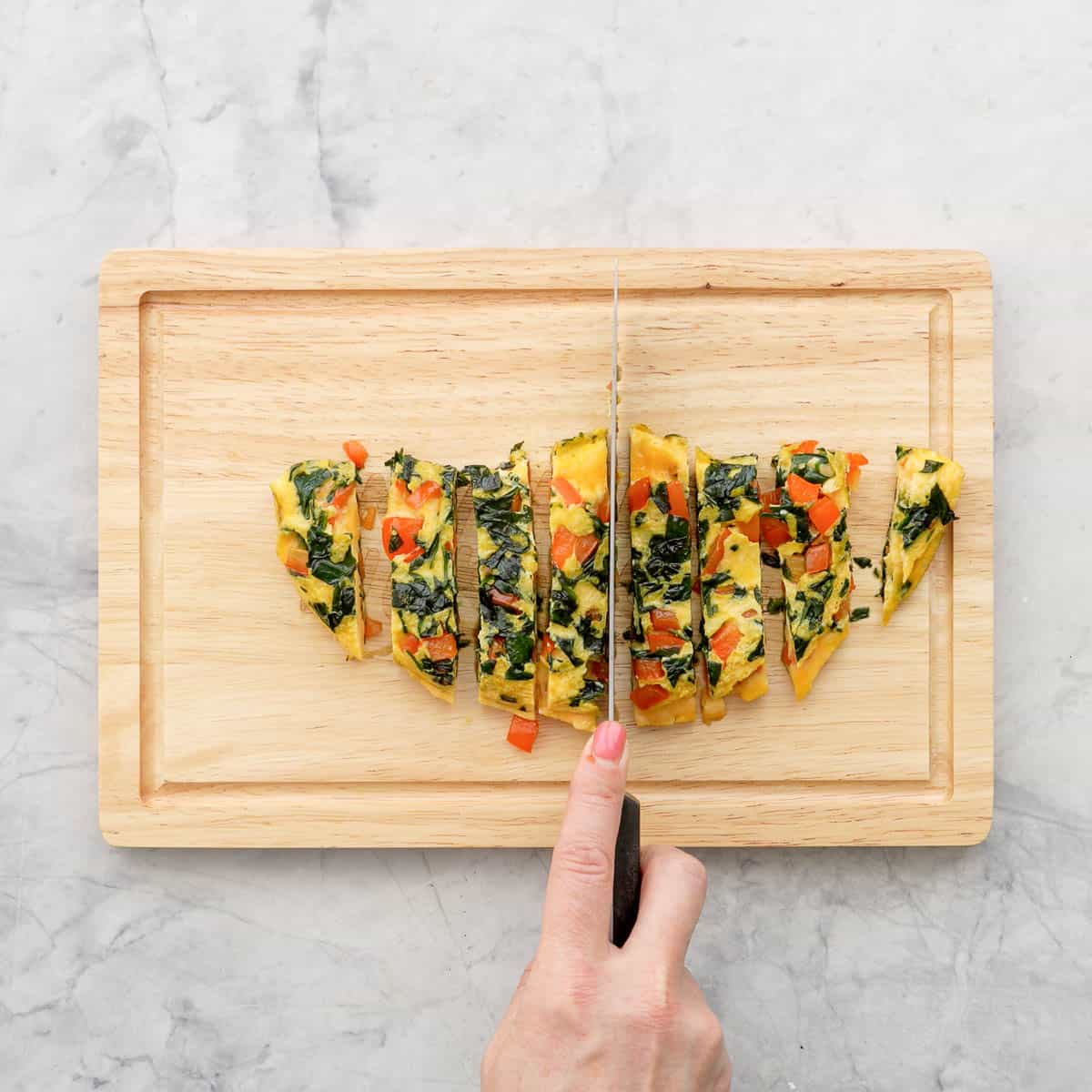 Spinach omelette folded in half on wooden chopping board being sliced with a knife into finger like strips.