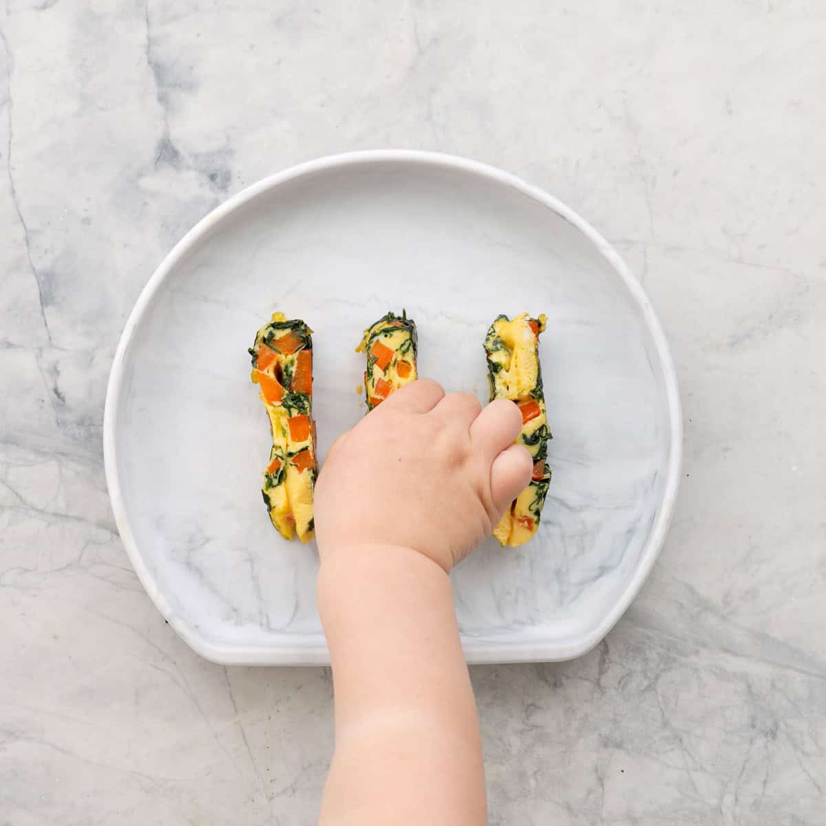 Three spinach omelette fingers on a child's silicone plate with a toddlers hand taking one of the fingers.