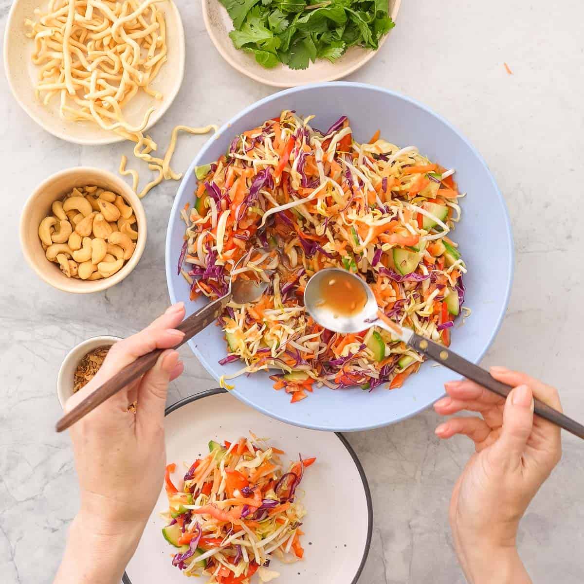 Large bowl of Asian Salad being served onto a dinner plate next to bowls with garnishes inside.