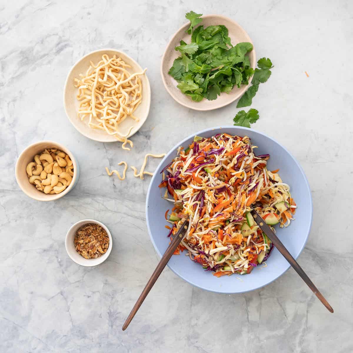 Large bowl of Asian Salad with utensils next to smaller bowls of garnishes on benchtop.