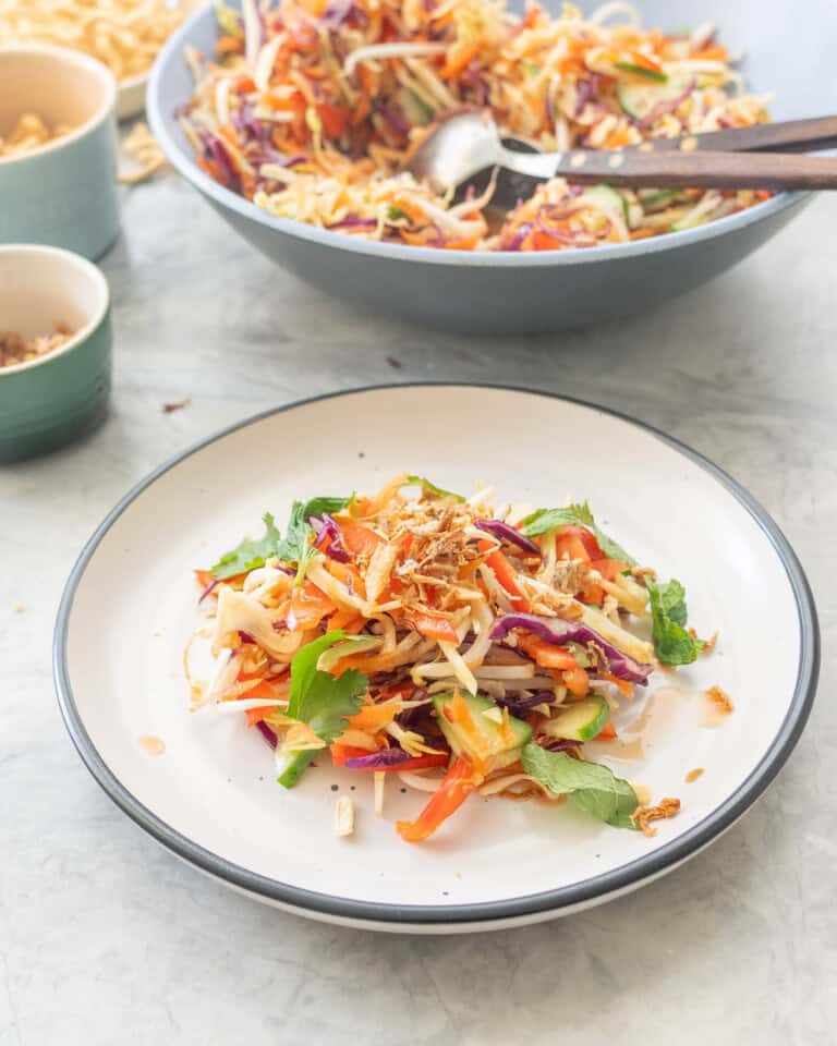 Asian Salad served on a dinner plate with garnish on top, Large bowl of asian salad in background and bowl of garnishes.