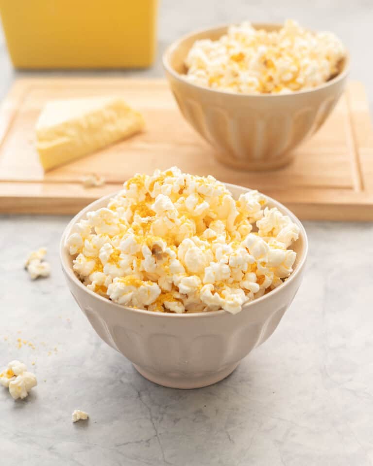 Cheese popcorn in a small bowl on bench top with block of cheese and bowl of cheese popcorn blurred in background on wooden chopping board.