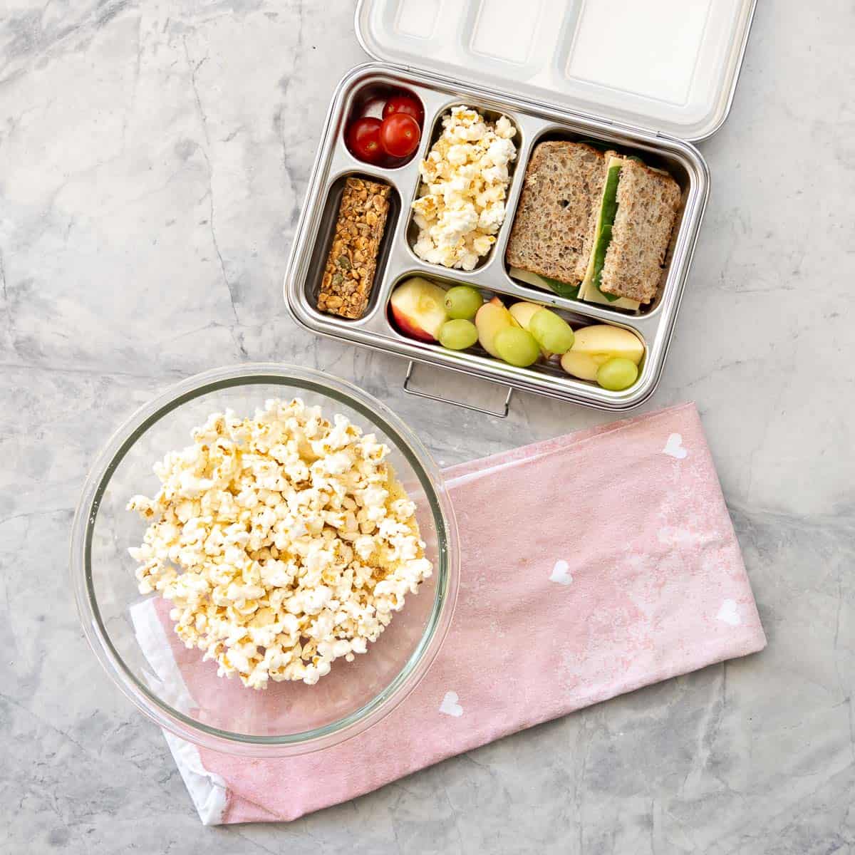 Cheese popcorn on benchtop in large glass bowl sitting on a pink tea towel, Childs lunchbox with Cheese popcorn, sandwich, apple, grapes, tomato and muesli bar inside.