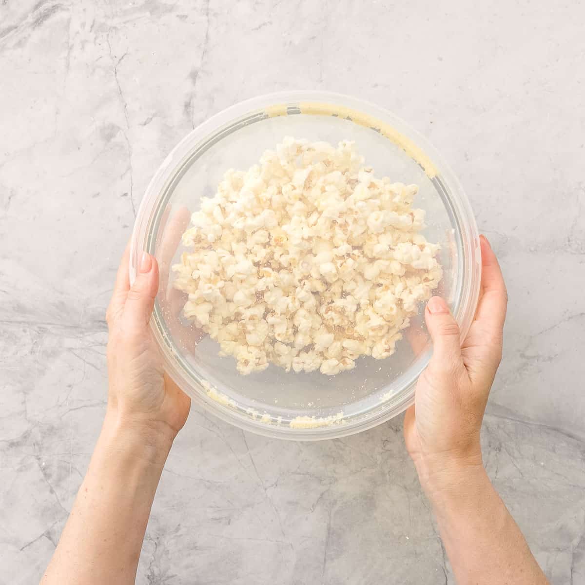 Hands holding a glass bowl sealed with clear lid on benchtop with popcorn and cheese inside.