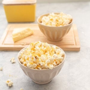 Cheese popcorn in a small bowl on bench top with block of cheese and bowl of cheese popcorn blurred in background on wooden chopping board.