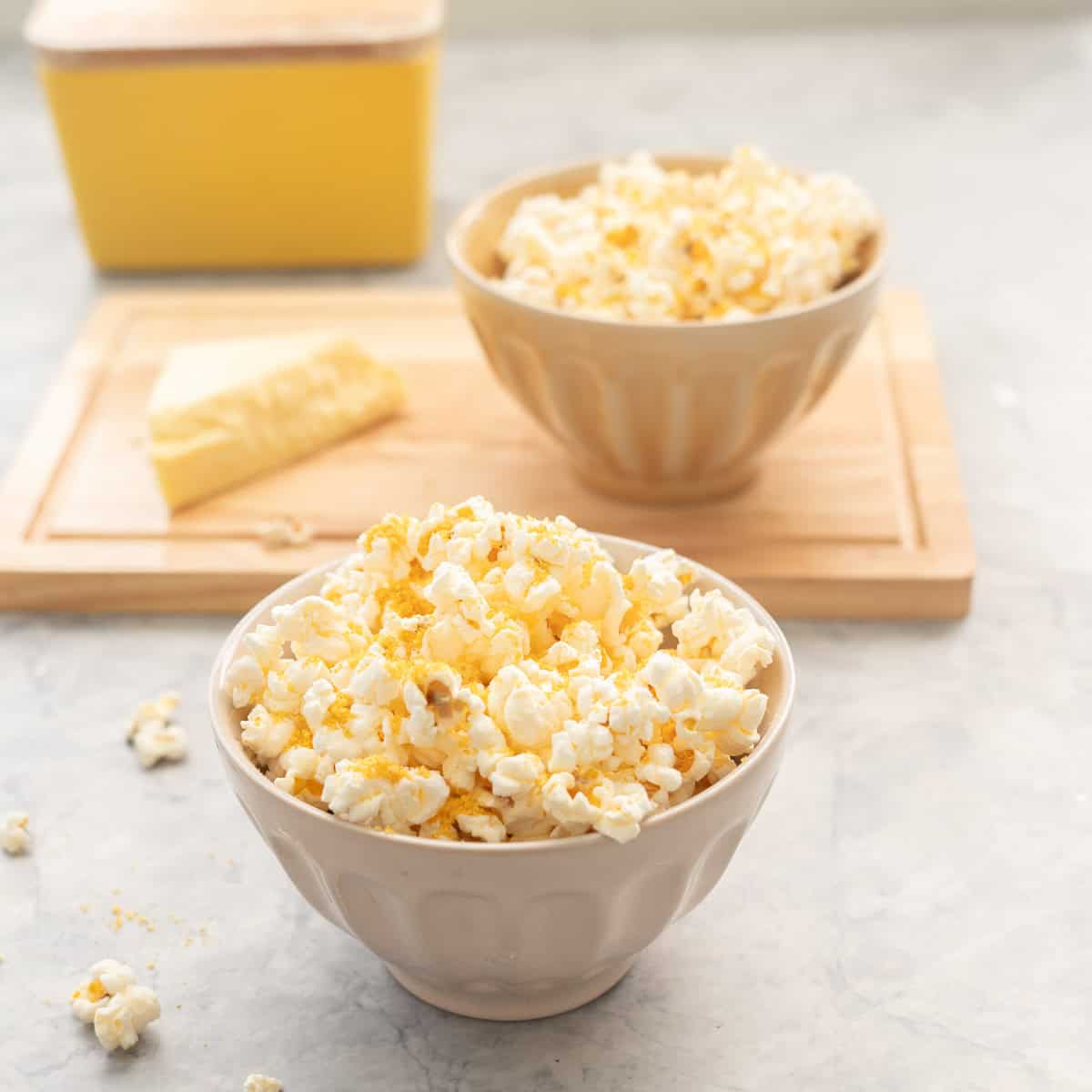 Cheese popcorn in a small bowl on bench top with block of cheese and bowl of cheese popcorn blurred in background on wooden chopping board.