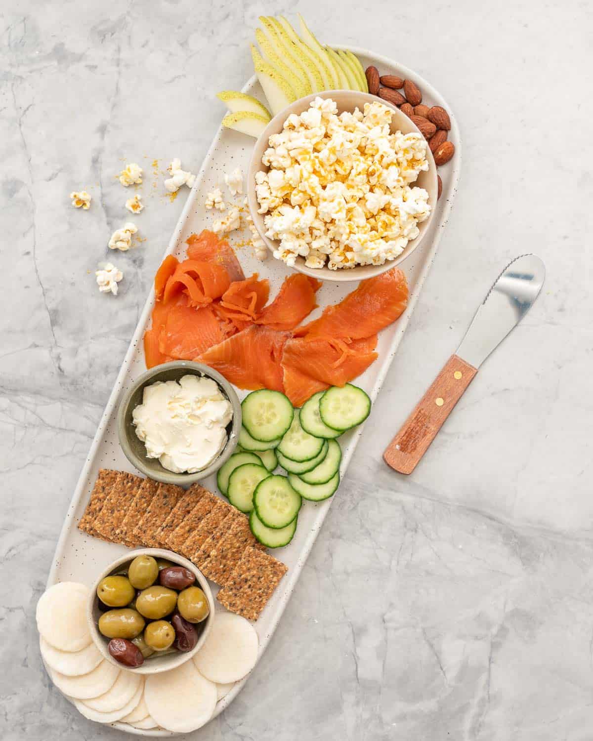 Cheese popcorn in bowl positioned on a long platter board on benchtop with variety of fresh foods- Cucumber, salmon, cream cheese, crackers, pear slices, almonds and olives.