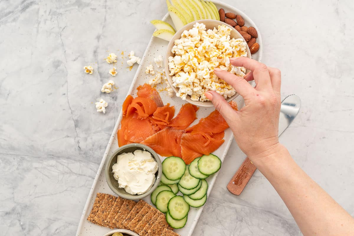 Hand taking a piece of Cheese popcorn from bowl positioned on a long platter board with variety of fresh foods, Cucumber, salmon, cream cheese, crackers, pear slices, almonds and olives.