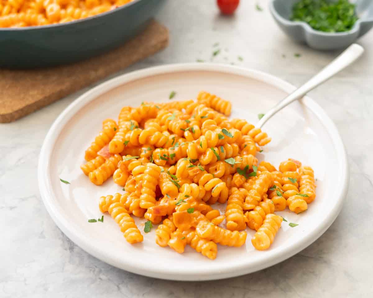 Portion of creamy tomato pasta served on plate garnished with fresh basil with skillet of remaining pasta, cherry tomatoes and small ramekin of basil in the background.