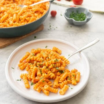 Portion of creamy tomato pasta served on plate garnished with fresh basil with skillet of remaining pasta, cherry tomatoes and small ramekin of basil in the background.