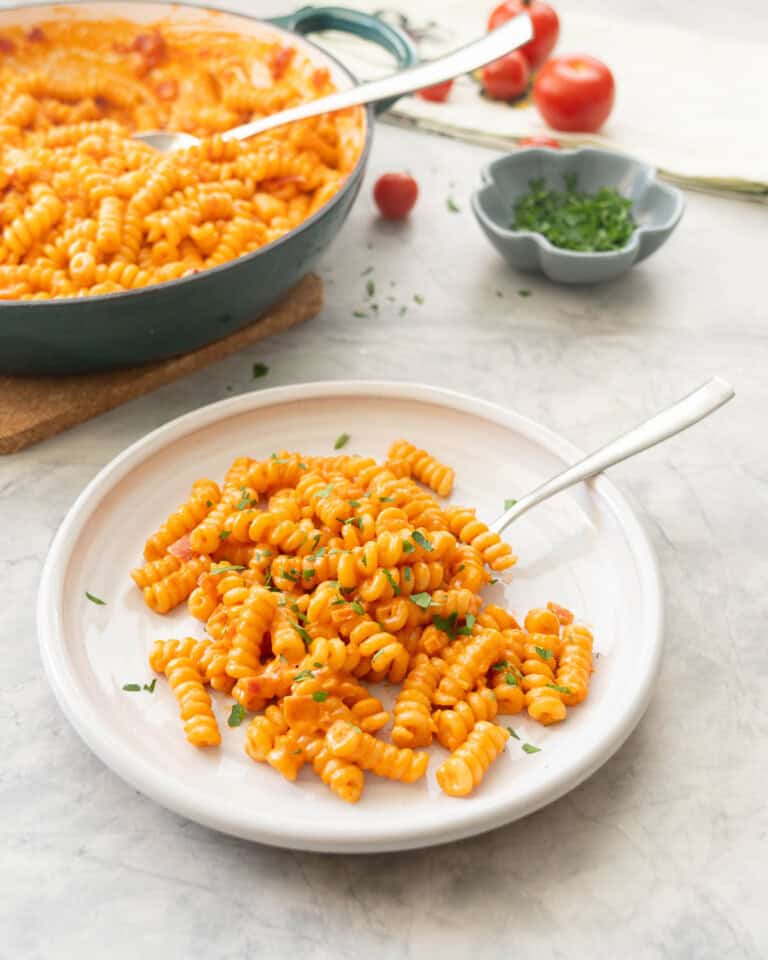 Portion of creamy tomato pasta served on plate garnished with fresh basil with skillet of remaining pasta, cherry tomatoes and small ramekin of basil in the background.