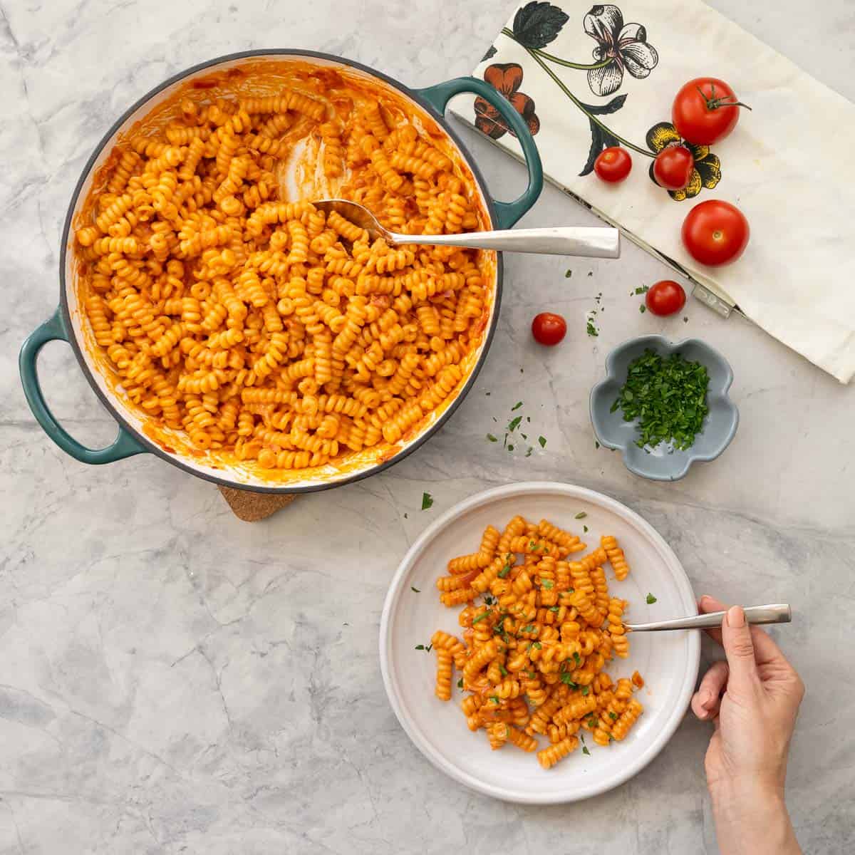 Portion of creamy tomato pasta served on plate with hand holding a fork, garnished with fresh basil with skillet of remaining pasta, cherry tomatoes and small ramekin of basil in the background.