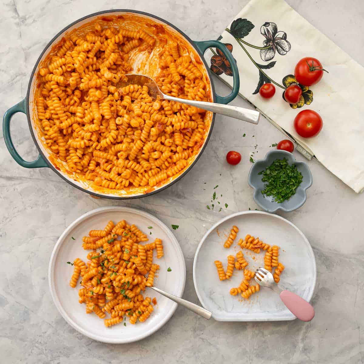 Portion of creamy tomato pasta served on plate garnished with fresh basil, a small serving on a silicone child plate and fork with skillet of remaining pasta, cherry tomatoes and small ramekin of basil in the background.