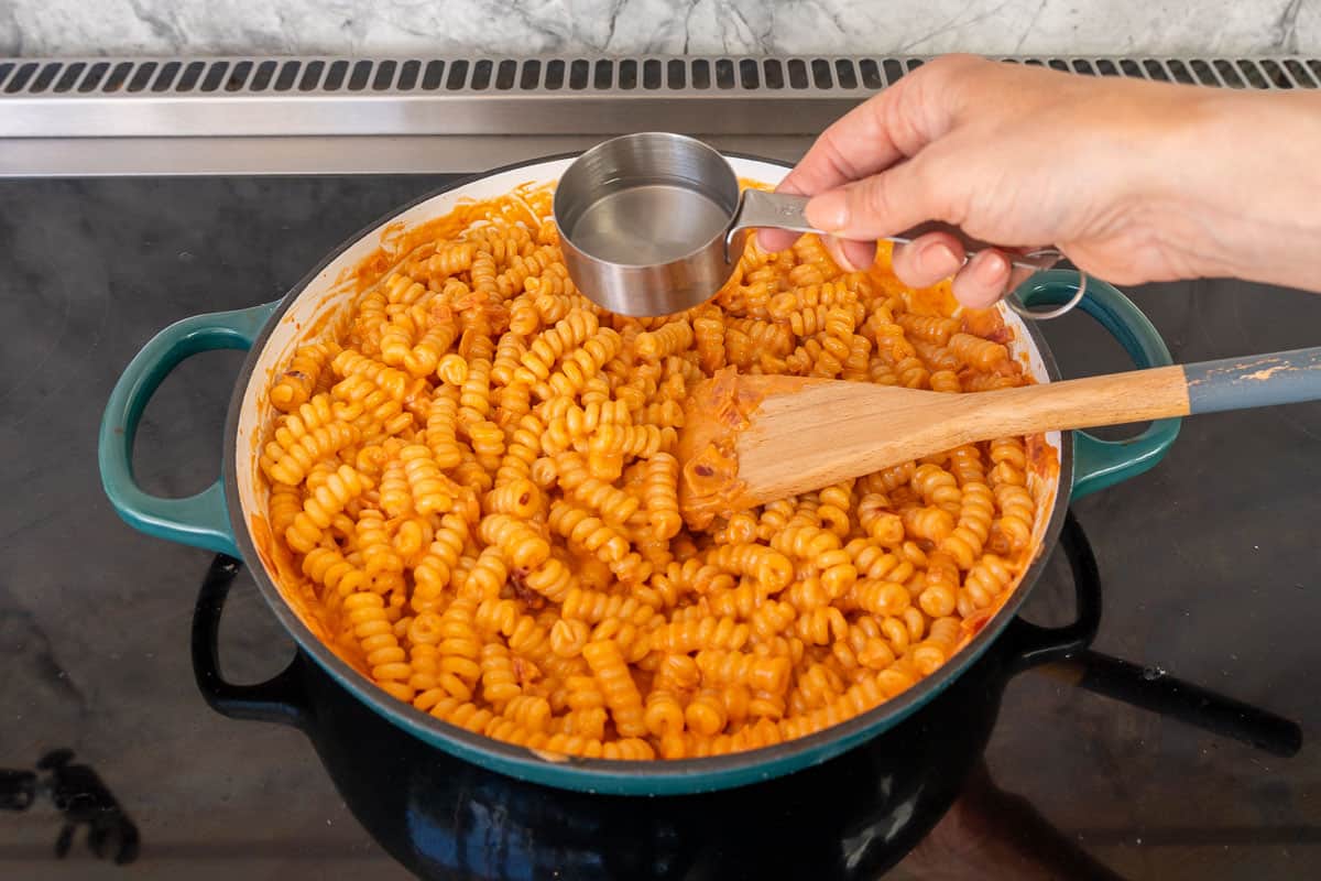 Creamy tomato pasta on stove top in skillet with hand holding measuring cup adding some pasta water in.